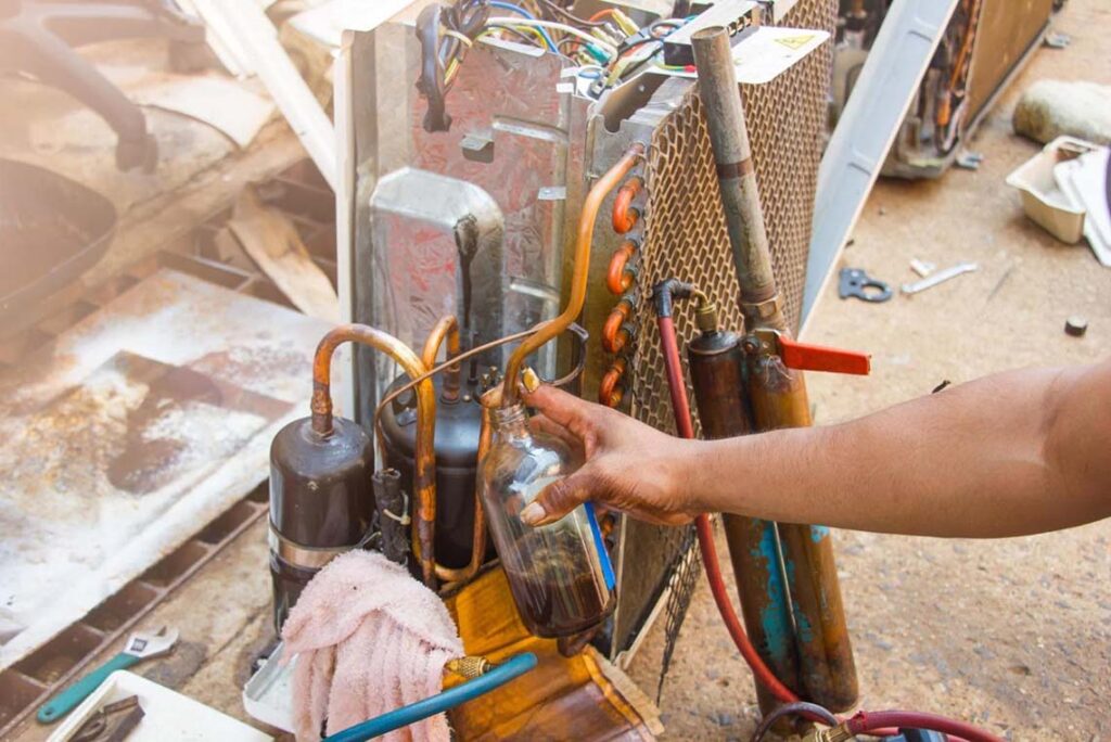 A close-up of an HVAC technician's hand draining fluid from an air conditioning unit, with exposed wiring and metal components visible under natural daylight.