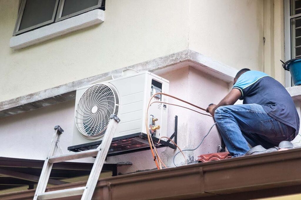 An HVAC technician installing an outdoor air conditioner compressor unit mounted on a wall.