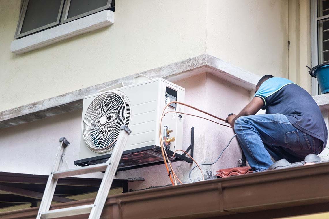 An HVAC technician installing an outdoor air conditioner compressor unit mounted on a wall.