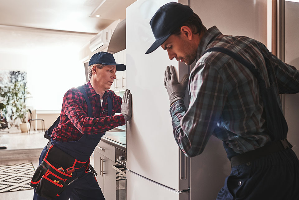 Two workers maneuver a large refrigerator in a modern kitchen, showcasing teamwork and careful handling of appliances.