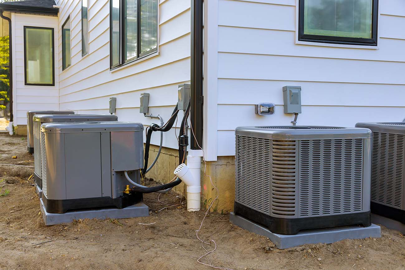Three gray outdoor HVAC units are installed alongside a white house, with pipes and electrical connections visible.