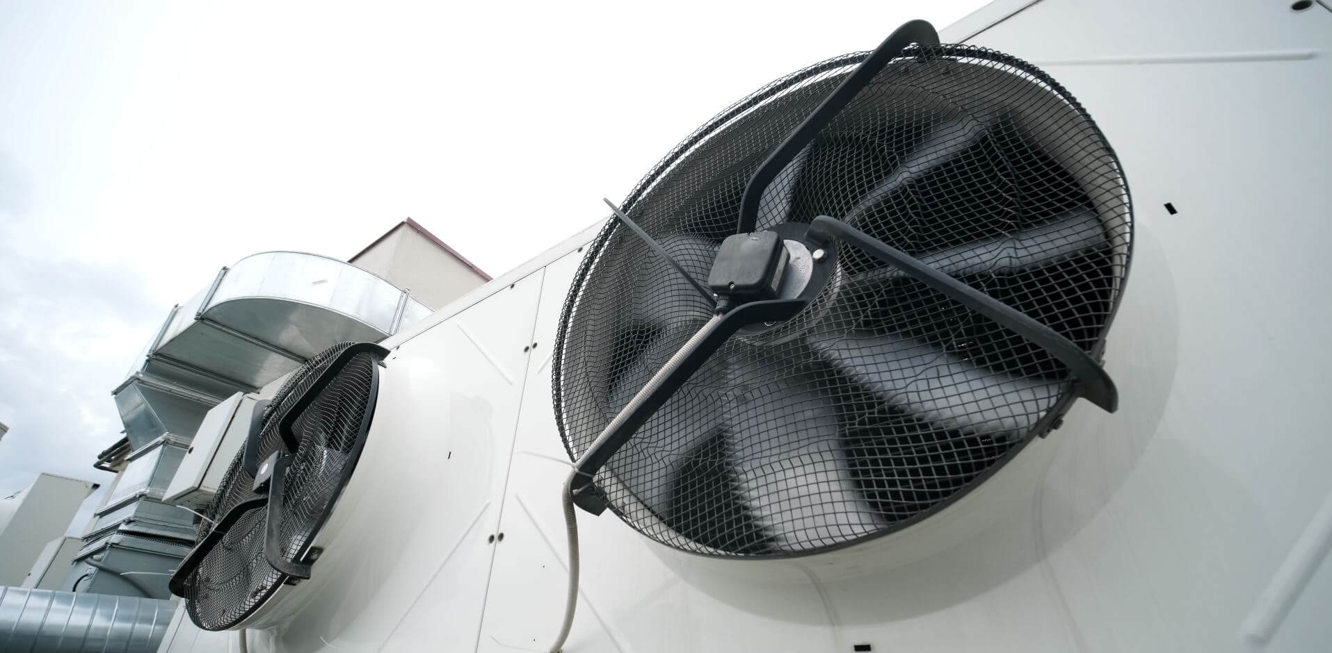 Two large industrial fans mounted on a white exterior wall, with ventilation ducts visible in the background under a cloudy sky.